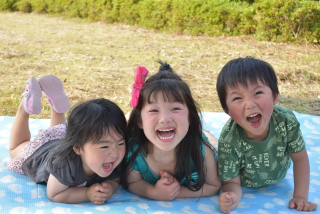 芝生に敷いたシートの上で、笑顔で寝転ぶ3人の子どもたち